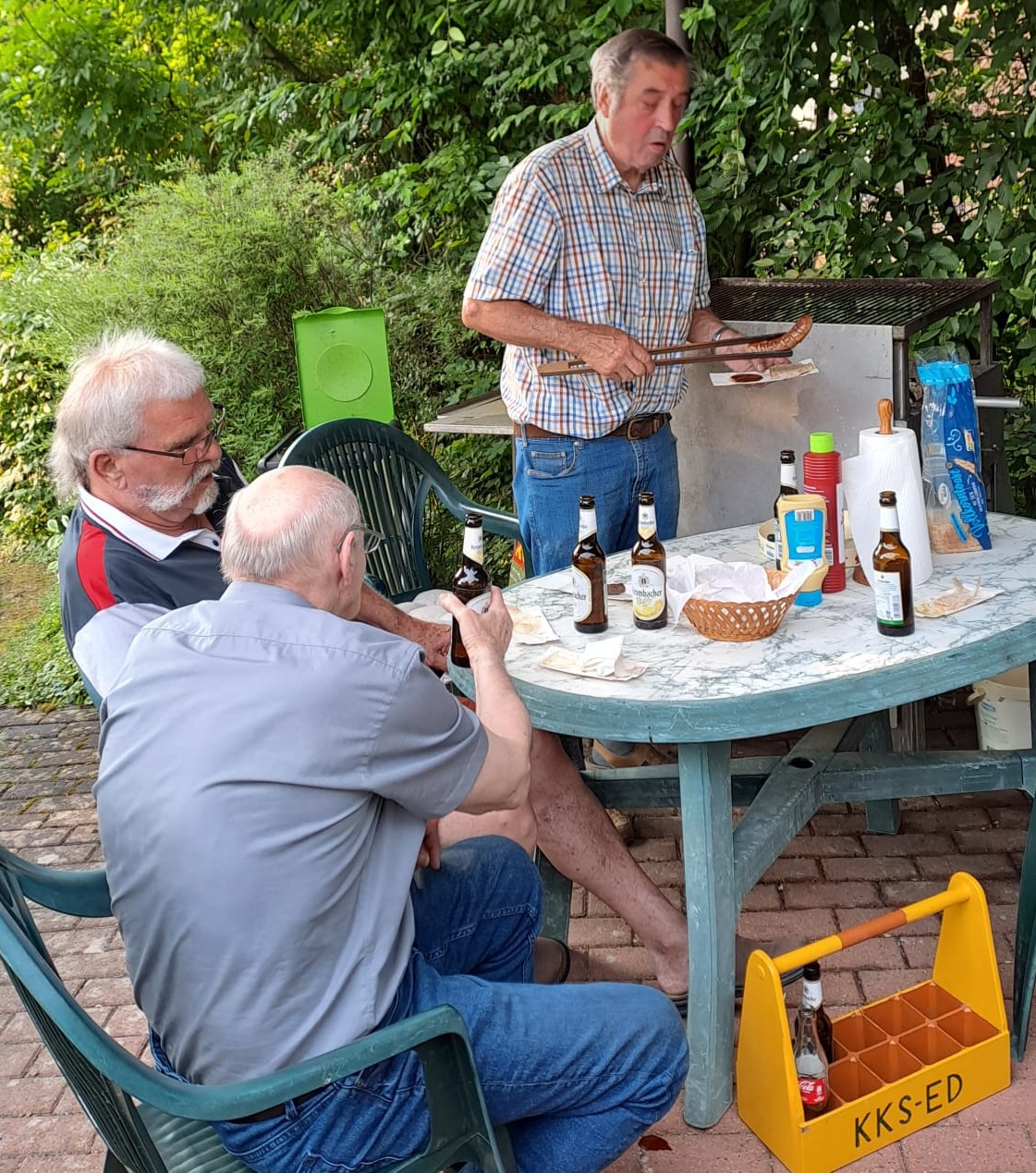 3 Männer sind auf dem Bild zu sehen, wobei ein Mann gerade eine gegrillte Wurst auftut. Sie sitzen draußen und auf dem Tisch stehen Bierflaschen, Ketchup und Brot.