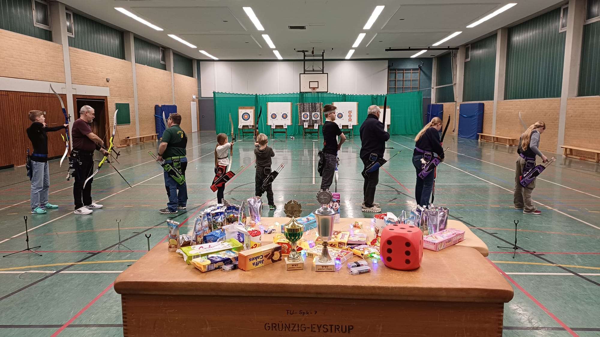 Auf dem Bild sind Bogenschützen und -schützinnen in einer Sporthalle. Sie haben ihren Bogen in der Hand und im Hintergrund sind die Scheiben aufgestellt. Im Vordergrund sind Süßigkeiten, Getränke und Deko zu sehen.