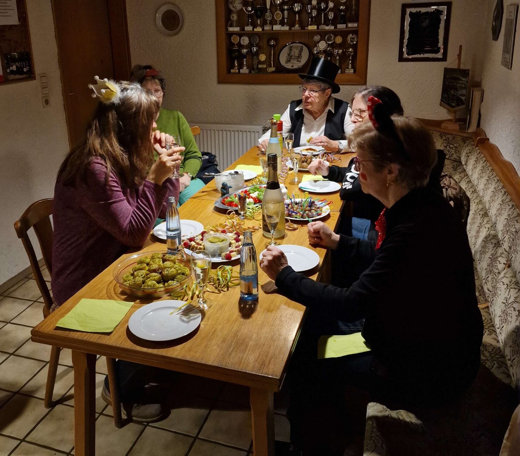 Auf dem Bild sitzen fünf Frauen zusammen um einen festlich gedeckten Tisch, wo selbstgemachtes Essen und Trinken draufsteht. Die Frauen sind passend zu Fasching verkleidet.