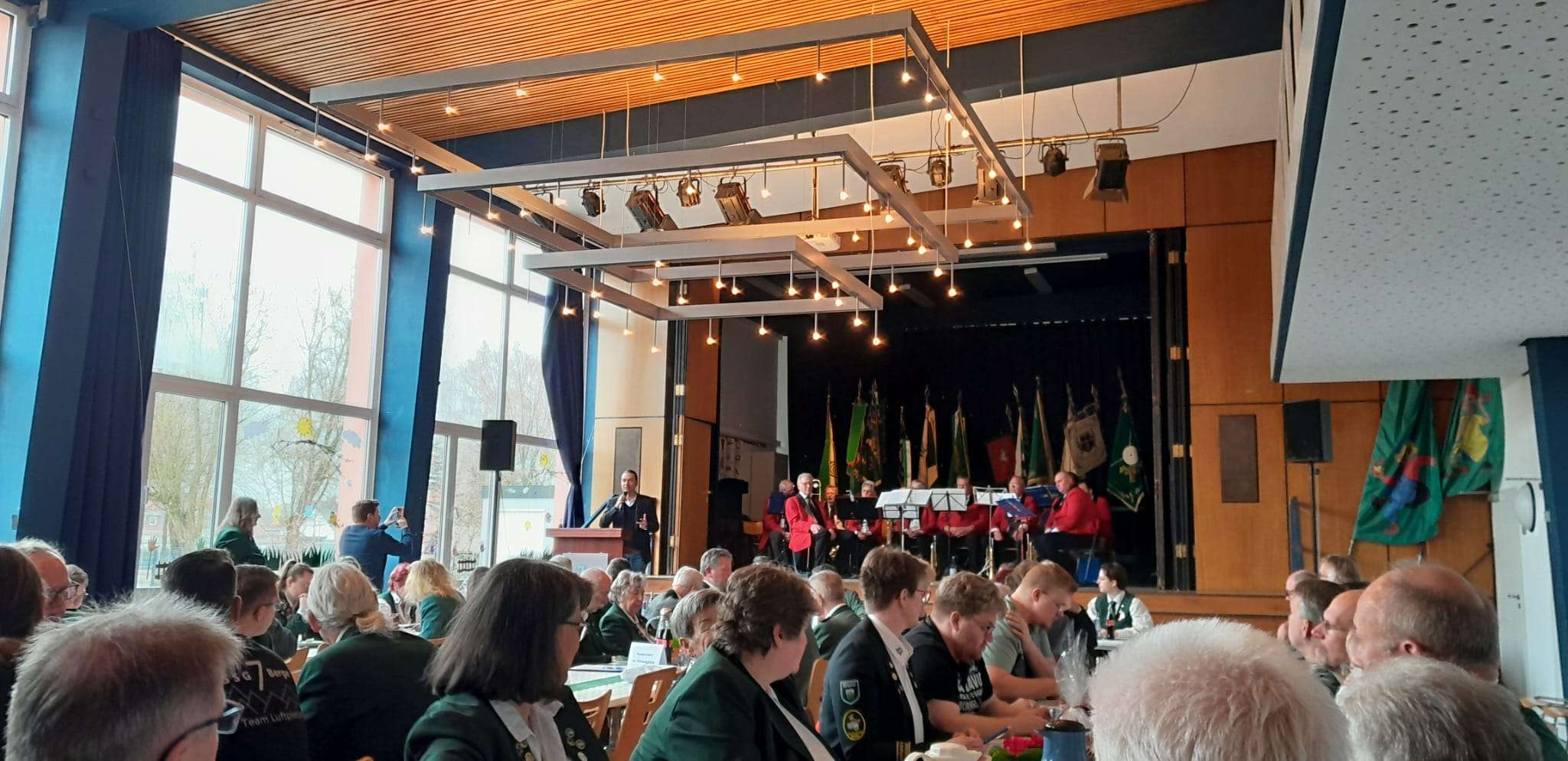 Auf dem Bild ist im Hintergrund eine Bühne zu sehen, auf der Musiker sitzen. Im Vordergrund sitzen viele Leute in Schützenuniform und schauen Richtung Bühne.