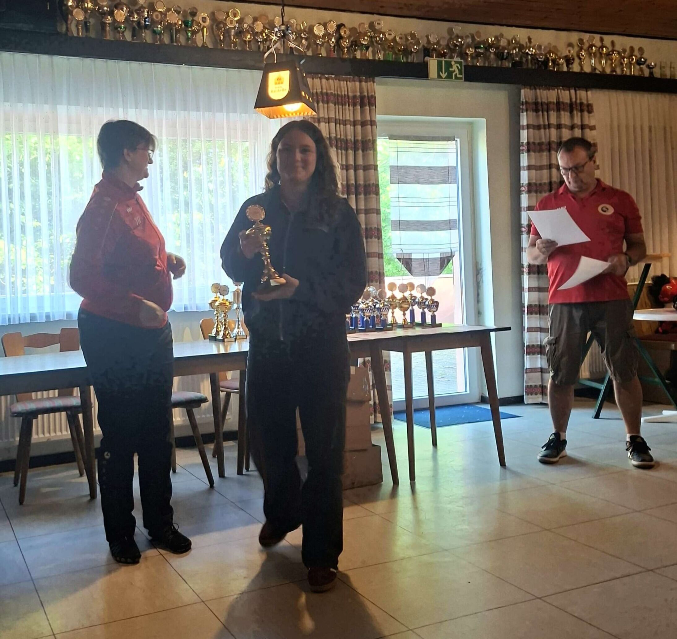 Auf dem Bild ist eine Jugendliche im Vordergrund zu sehen, die ein Pokal in beiden Händen hält. Im Hintergrund sind zwei weitere Personen in Vereinsklamotten und es stehen Pokale auf Tischen.