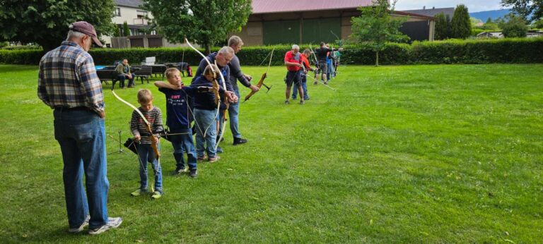 Es sind Jugendliche und Erwachsene auf einer Wiese zu sehen, die mit Pfeil und Bogen trainieren. Sie stehen in einer Reihe.