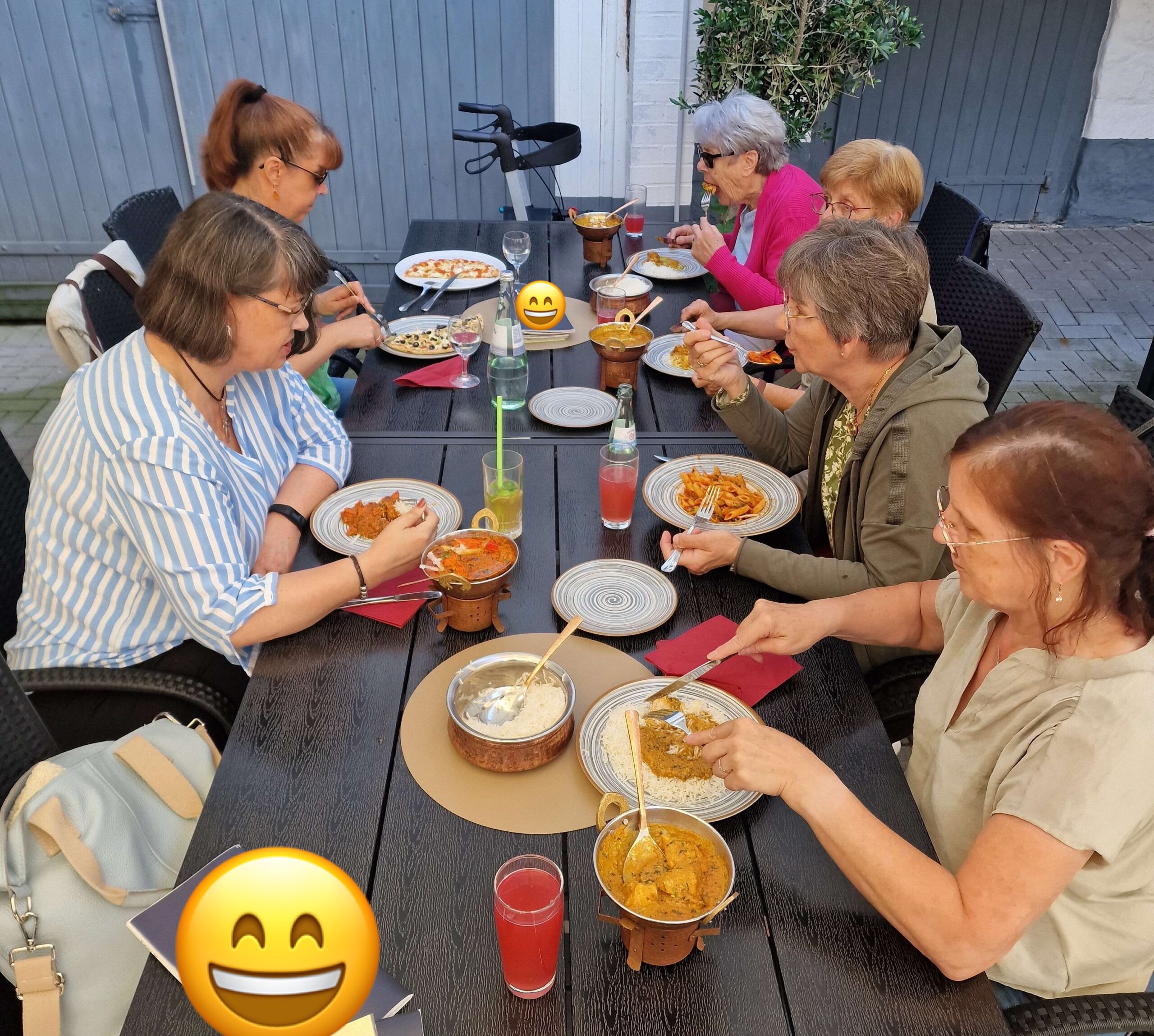 Auf dem Bild sitzen draußen 6 Frauen an einem Tisch mit warmen Speisen und Getränken und essen gemeinsam