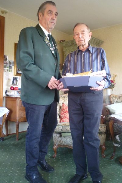 Auf dem Bild sind zwei Männer zu sehen. Der linke Mann hat eine Schützen-Uniform an. Der rechte Mann hält ein Geschenkkorb in der Hand. Beide schauen in die Kamera.